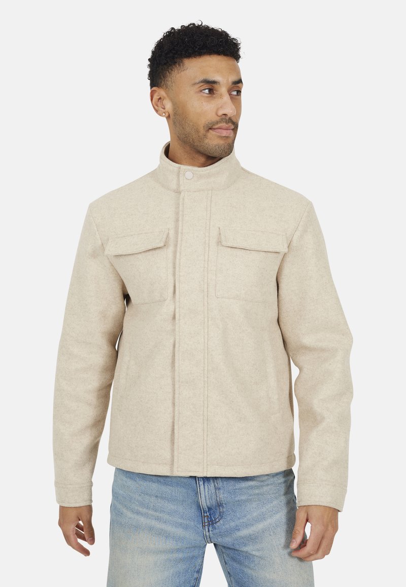 Veste en laine beige avec col montant, deux poches à rabat à l'avant et coupe droite. Associée à un jean en denim bleu. Texture lisse et structurée.