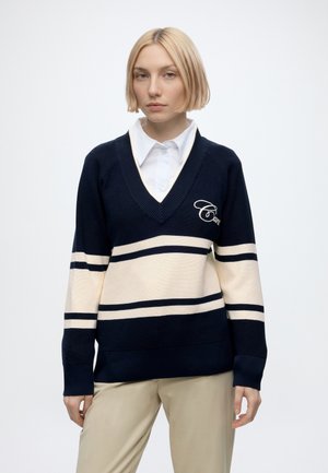 Junge Frau mit kurzem blondem Haar, die einen marineblau-creme gestreiften Pullover über einem weißen Hemd mit Kragen und beigen Hosen trägt.