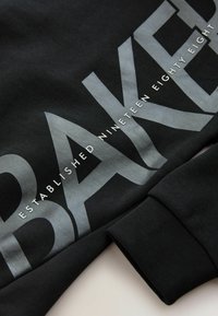 Fekete pulóver nagy ezüst betűkkel, amelyeken a "BAKER" felirat olvasható, valamint kisebb fehér szöveg: "ESTABLISHED NINETEEN EIGHTY EIGHT". Puha tapintású, bordázott mandzsettákkal.