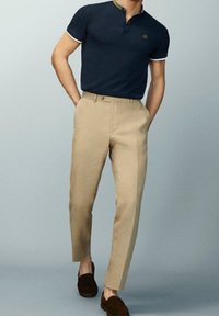 Mies, joka pukeutunut tummansiniseen lyhythihaiseen polo-paitaan, beigeihin muotoiltuihin housuihin ja tummanruskeisiin loafereihin, seisoo kädet taskuissa.