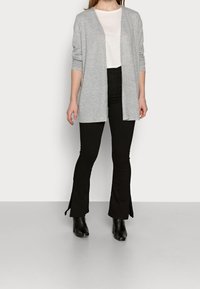 Cardigan gris claro, abierto en el frente, con mangas largas, usado sobre una blusa blanca y pantalones negros acampanados. Botas de tacón negras complementan el conjunto.