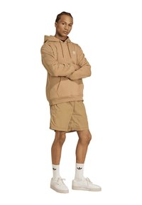 Lichtbruine hoodie met trekkoorden, beige korte broek en witte sneakers. Model staat met gekruiste armen en toont casual sportkleding.