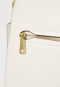 Cream leather bag close-up featuring a gold zipper and tag. The zipper pull is engraved with the brand name "COACH." Smooth texture.