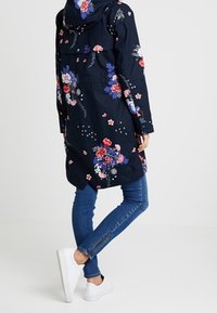 Mørkeblå blomstrede jakke med hætte, der har et farverigt blomstermønster og en løs pasform. Sammen med mørkeblå jeans og hvide sneakers.
