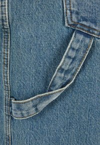 Material denim de culoare albastru deschis cu suprafață texturată; prezintă o clapetă de buzunar cusută și cusături detaliate portocalii de-a lungul marginilor.