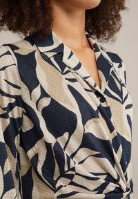 Hemd mit kragenförmigem Ausschnitt in Marineblau und Beige, aus strukturiertem Stoff und mit einem abstrakten Blattmuster. Knopfdetail am Kragen sichtbar.