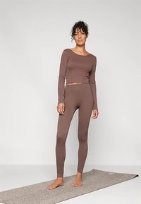 Rudas, liesas ilgomis rankovėmis viršus ir derančios tamprės iš tamprios medžiagos, eksponuojami ant jogos kilimėlio. Lygi tekstūra, minimalistinis dizainas.