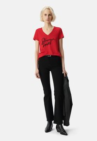Camiseta roja de cuello en V con la frase "Stronger Love" en cursiva negra, combinada con unos vaqueros negros de campana y botas de tobillo negras, sosteniendo una chaqueta oscura.