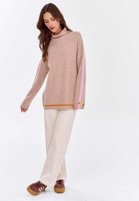Hellbrauner Pullover mit rosa und orangen Akzenten, ausgestattet mit einem hohen Kragen, langen Ärmeln und einem lässigen Schnitt. Kombiniert mit cremefarbenen Hosen und braunen Sneakers.