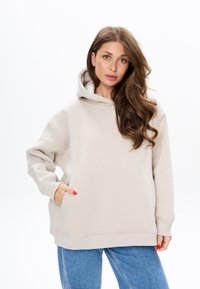 Hoodie oversize beige chiaro realizzato in tessuto morbido, con tasca frontale, cappuccio con coulisse e polsini e orlo a costine. Abbinato a jeans blu.