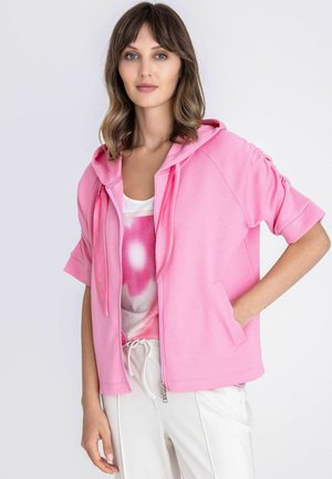 Vrouw draagt een lichtroze korte mouwen zip hoodie over een wit en roze tanktop en witte broek met trekkoord, staand met één hand in de zak.