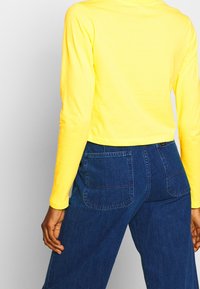 Gul långärmad kort topp i kombination med mörkblå denimbyxor med två bakfickor och synliga sömmar.