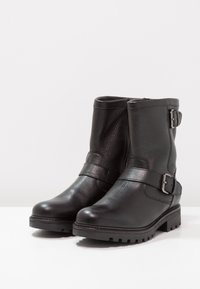 Botas de cuero negro hasta el tobillo con un acabado texturizado, que cuentan con un detalle de correa y hebilla, suela de goma resistente y punta redondeada.