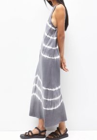 Šedé tie-dye maxi šaty s bílými horizontálními pruhy, s bezrukávovým designem a volným střihem. Kombinováno s černými tkanými sandály.