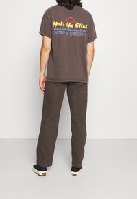 Hnědé bavlněné tričko s grafickým textem a designem hor na zádech. Kombinováno s tmavě hnědými kalhotami a černými botami. Minimalistický styl.