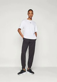 Kvinne står med hendene i lommene iført hvit Puma t-skjorte, svarte joggebukser og svarte joggesko mot en lys, ensfarget bakgrunn.