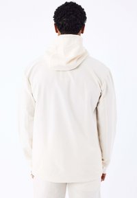 Man in een off-white hoodie en bijpassende broek, gezien vanaf de achterkant tegen een eenvoudige witte achtergrond.
