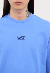 Modrá mikina vyrobená z jemnej textílie, s okrúhlym výstrihom a čiernym logom EA7 Emporio Armani na hrudi. Jednoduchý, neformálny dizajn.