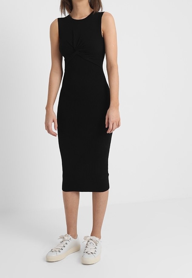 Femme portant une robe midi noire sans manches avec un détail torsadé à l'avant, associée à des baskets blanches à lacets, debout devant un fond blanc uni.