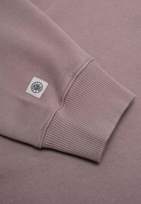 Vaaleanpunainen collegepaita, jossa on resorit. Siinä on pieni valkoinen kudottu etiketti mustalla tekstillä. Sujuva pinta ja rento muotoilu.