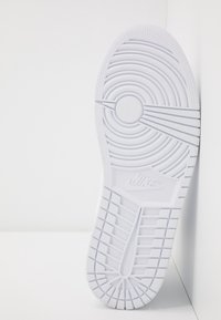 Semelle d’une basket Nike blanche avec des motifs de bande de roulement circulaires et rectangulaires, placée verticalement contre un mur blanc.