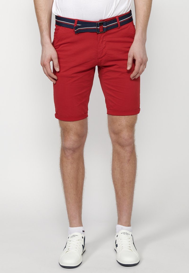 Shorts en coton rouge avec des ourlets retroussés, dotés d'une ceinture rayée marine. Associés à des baskets blanches et un t-shirt blanc.