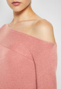 Pull en maille rose clair avec un design décolleté. Tissu texturé et encolure large. Texture douce, silhouette ajustée et finition élégante.