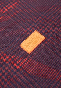 Gestructureerde stof met een rood en marineblauw houndstooth patroon; heeft een kenmerkend oranje rubberen patch met reliëfbranding en details.