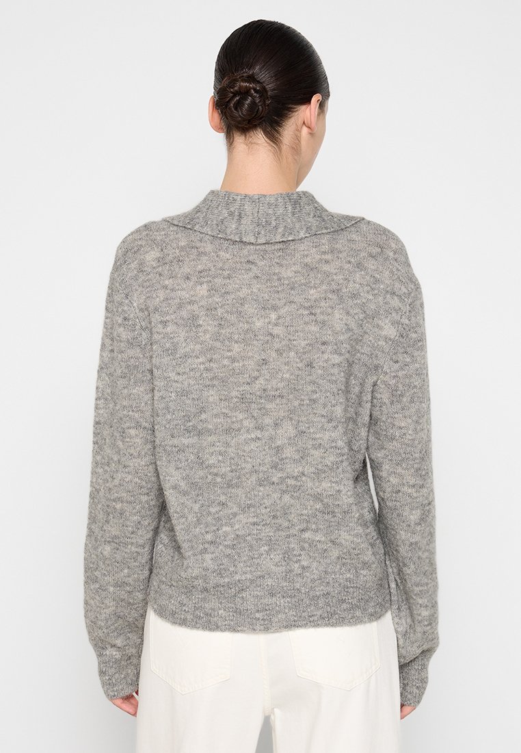 Szary sweter z dzianiny o fakturowanym wykończeniu, z szerokim kołnierzem i opadającymi ramionami, noszony na beżowych spodniach.