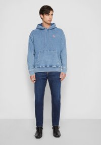 Light blue denim hoodie with kangaroo pocket, drawstring hood, and logo. Paired with dark blue jeans and black boots, standing on a plain background.