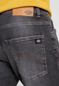 Takapocket ja vyötärö mustissa farkuissa, joissa näkyvät Dickies-merkin nahka- ja kangaslabelit, yhdistettynä keltaiseen paitaan.