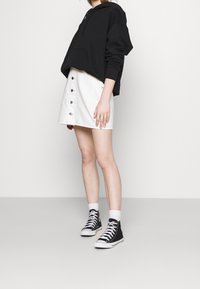 Felpa nera corta, gonna in denim bianca con bottoni, e sneaker alte nere con suole bianche. Il modello è in piedi su uno sfondo semplice.