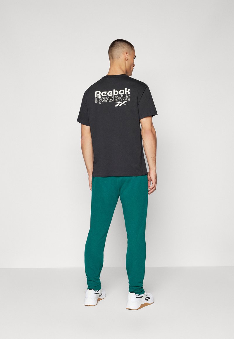 Camiseta negra con el logo blanco de "Reebok" en la parte posterior, combinada con pantalones de chándal turquesa y zapatillas blancas. Ropa deportiva simple y casual.