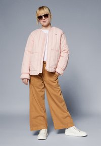 Rosa quilted jakke med frontlommer, kombinert med brede brune bukser og hvite sneakers. Modellen har på seg mørke solbriller og en hvit skjorte.
