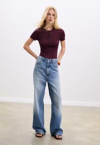 Bordeauxfarbener, taillierter Oberteil mit kurzen Ärmeln, kombiniert mit weit geschnittenen hellblauen Jeans. Bemerkenswerte Details: hohe Taille, ausgewaschene Textur und Kontrastnähte.