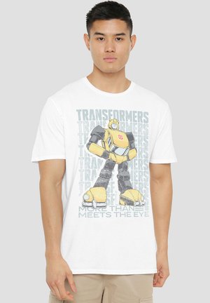 Weißes T-Shirt mit einem gelben Robotermotiv und schwarzen Akzenten, Text "Transformers" in Grau und dem Satz "Mehr als das Auge sieht."