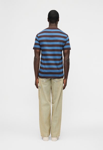 Άνδρας με την πλάτη προς την κάμερα, φοράει μπλε και καφέ ριγωτό μπλουζάκι, ανοιχτό μπεζ παντελόνι με φαρδιά μπάρα και λευκά αθλητικά παπούτσια.