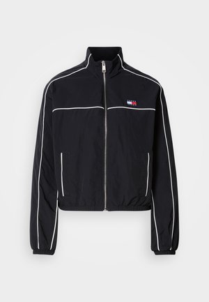 Veste noire courte avec passepoil blanc, dotée d'un col montant, d'une fermeture éclair frontale et d'un petit logo sur la poitrine. Fabriquée en tissu léger.