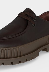 Chaussure en cuir marron foncé avec une semelle en caoutchouc épaisse et texturée, et des lacets marron assortis croisant deux œillets métalliques.