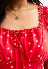 Rotes Kleid mit Punkten, das über einen quadratischen Ausschnitt, eine Kordelzugdetails an der Vorderseite und Puffärmeln verfügt. Sichtbare goldene, gestapelte Halsketten.