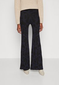 Pantaloni evazați cu talie înaltă, de culoare albastru închis, cu un model texturat. Materialul are un ușor luciu și o finisare netedă. Purtați cu un pulover bej.