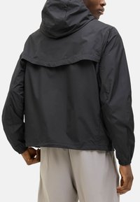 Veste noire en nylon avec capuche, dotée de poignets élastiques, d'une coupe ample et d'un design de dos superposé. Le tissu a une texture lisse et mate.