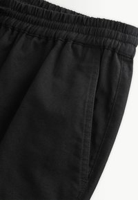 Pantalon en tissu noir avec une ceinture élastique et une poche latérale avec des coutures visibles sur un fond blanc.