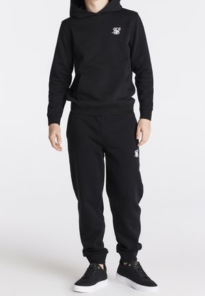 Sudadera negra con capucha y bolsillo canguro, con logo en el pecho, combinada con pantalones jogger negros con puños elásticos y zapatillas negras a juego.