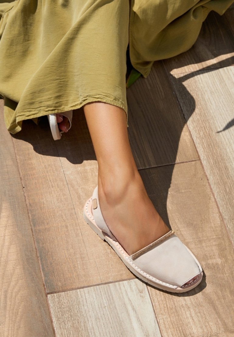 Zapato beige sin cordones con punta redonda, material de ante suave, costuras en contraste y acento metálico en la abertura, descansando sobre un suelo de madera.