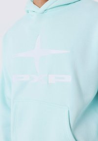 Haut en coton bleu clair avec une poche kangourou, logo blanc avec une étoile et le texte "PXP", et des poignets côtelés. Texture lisse.