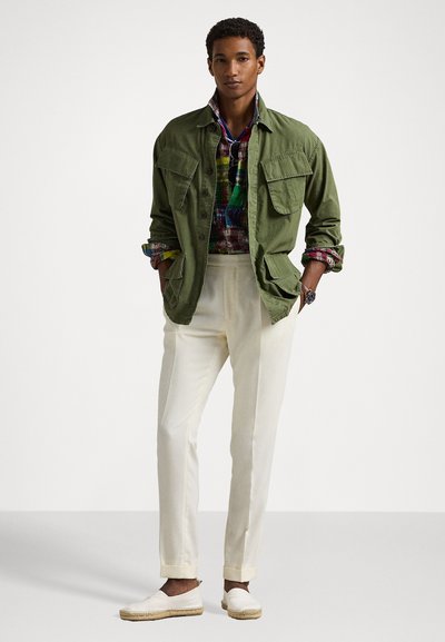 Casaco utilitário verde sobre uma camisa de xadrez multicolorida, calças brancas bem cortadas e espadrilhas brancas, com bolsos e mangas dobradas.