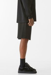 Crna majica dugih rukava uparena sa crnim kratkim hlačama do koljena. Crne atletske cipele s reljefnim naglaskom i rebraste čarape upotpunjuju odjevnu kombinaciju.