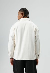 Witte fleecejas met kraag, knoopmanchetten en een comfortabele pasvorm, gecombineerd met donkere broek, gezien van achteren.