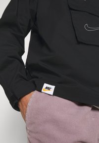 Person wearing a black Nike jacket with logo patch near the hem and hand in light purple corduroy pants pocket.